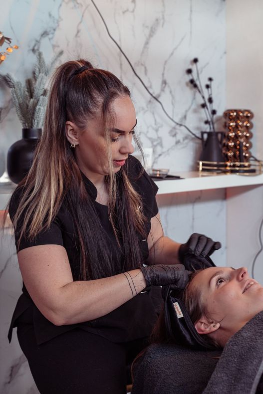 Een schoonheidsspecialist is bezig met het haar van een klant in een salon, met een marmeren muur en planken op de achtergrond.