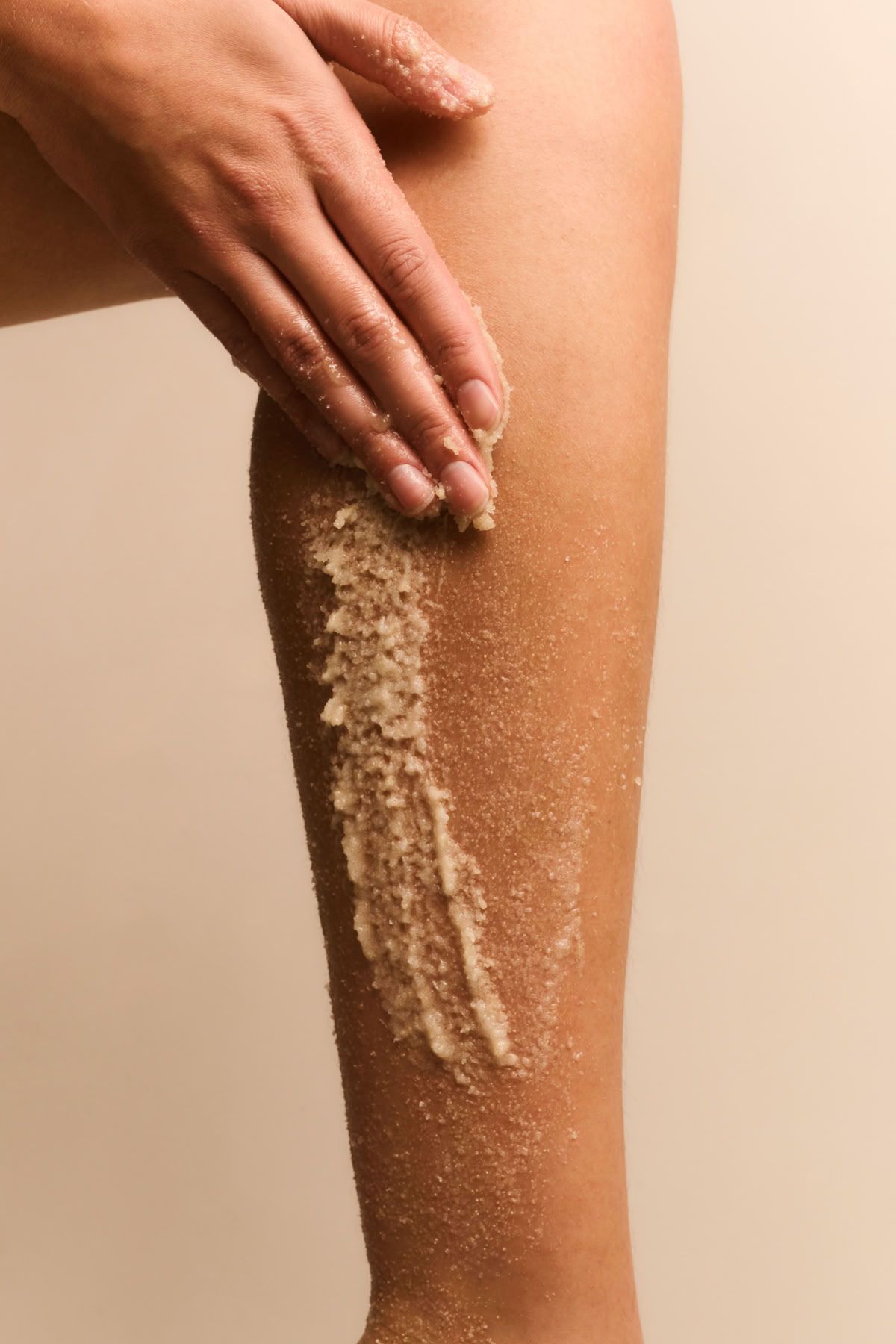 Hand die een exfoliërende scrub aanbrengt op een been, tegen een beige achtergrond.