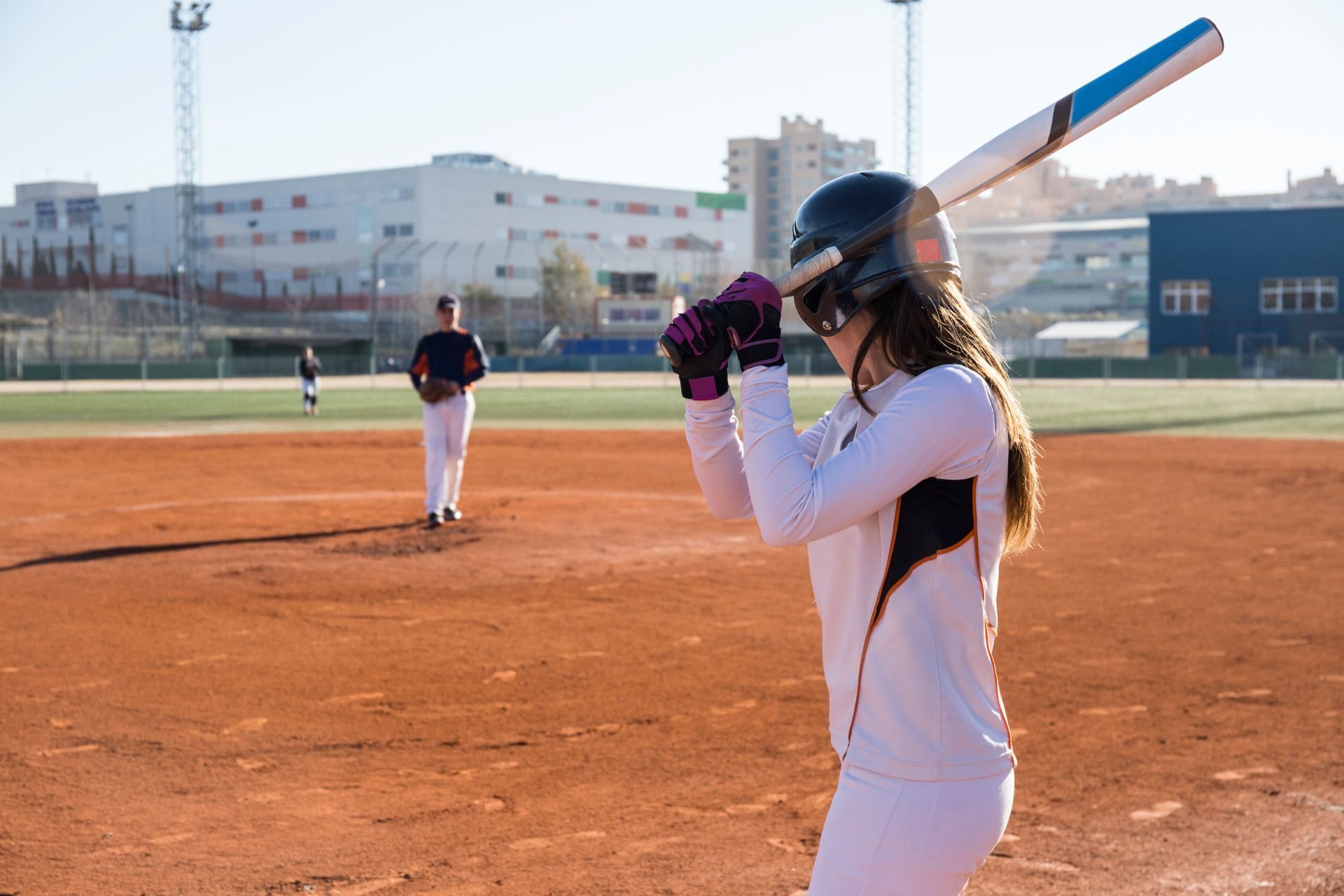 Baseball player in white uniform preparing to bat on a sunny field with a pitcher in the background
