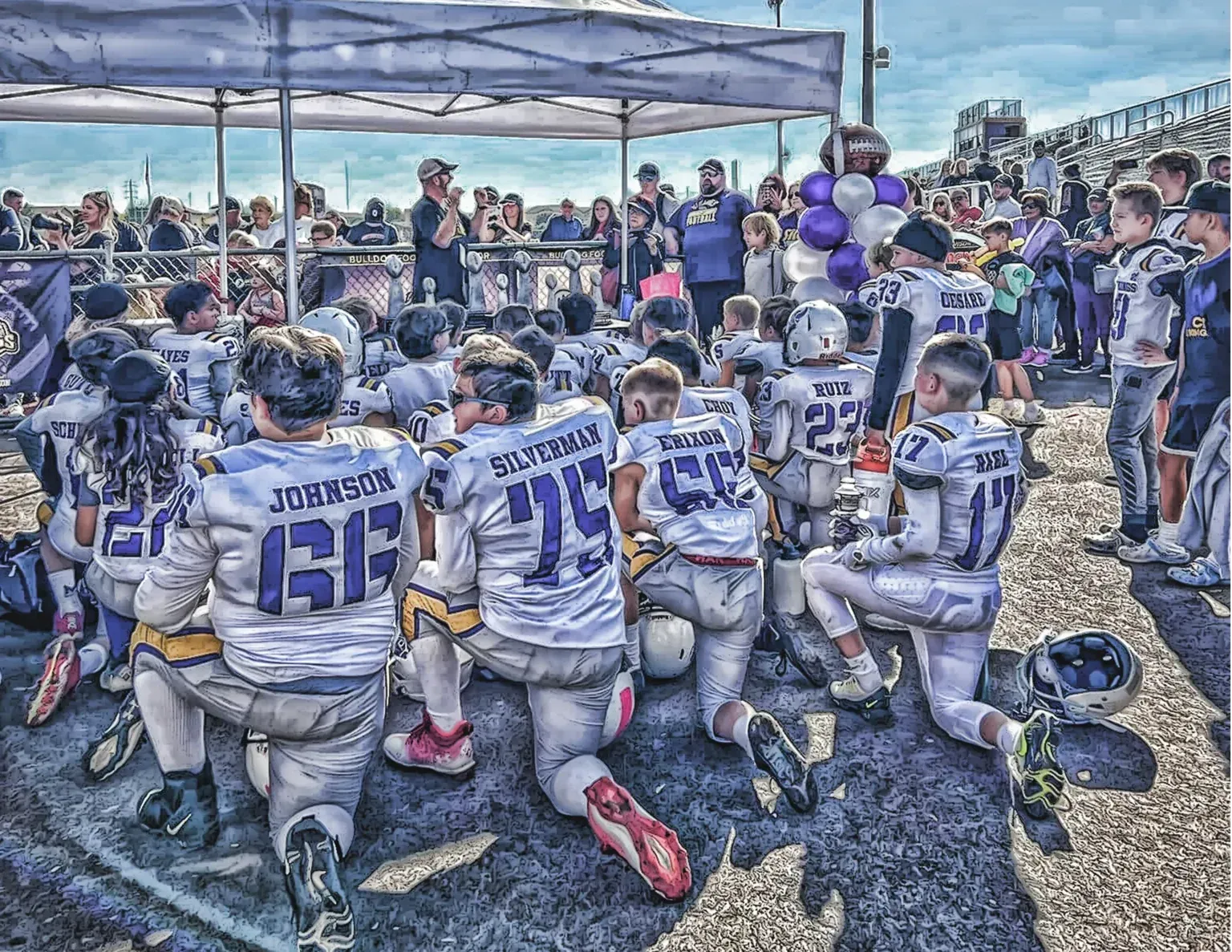 Football team kneeling in gray uniforms on a field, facing a cheering crowd under a canopy.