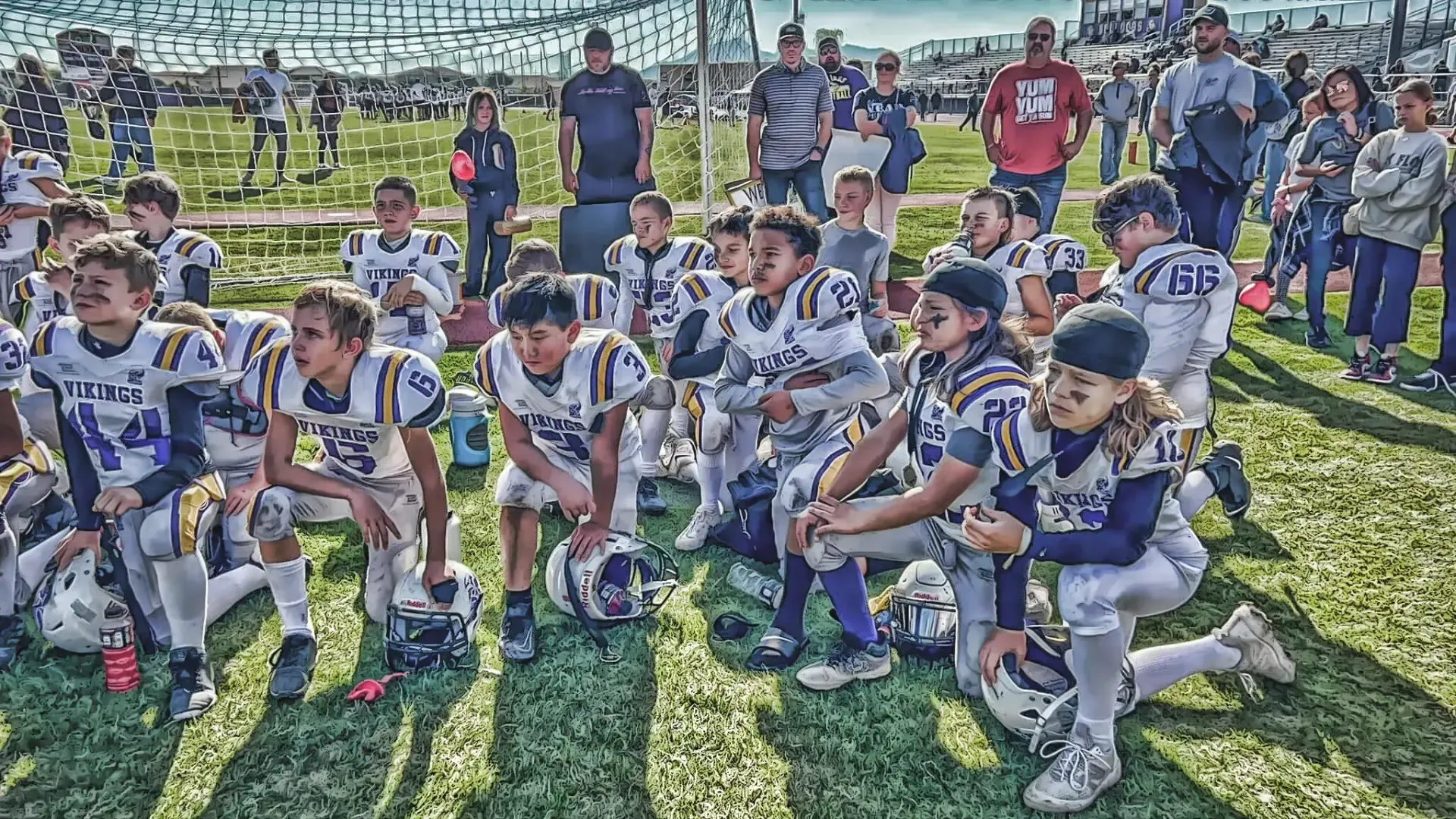 Football players in white and gray uniforms sitting on grass during a team meeting outdoors