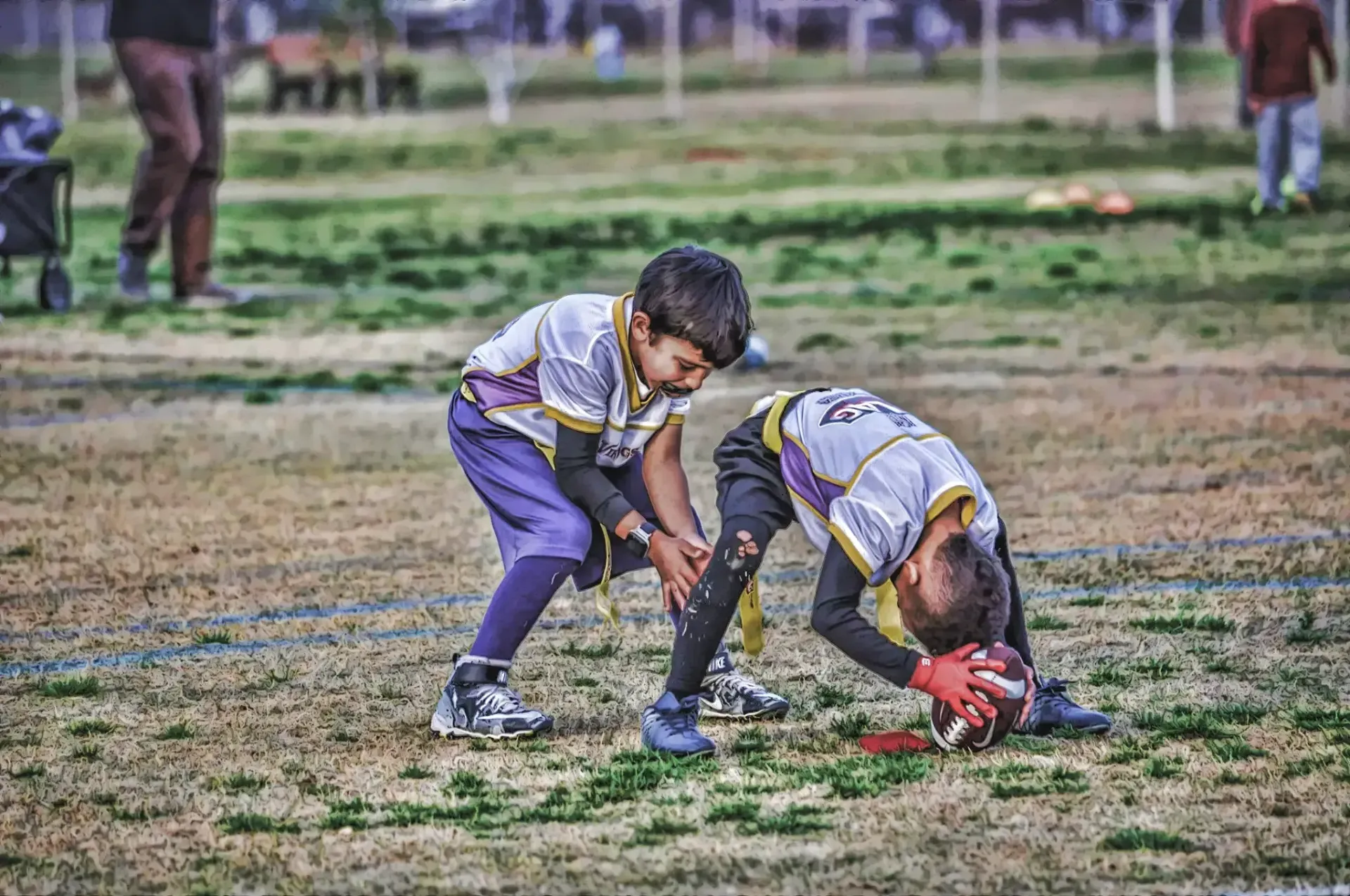 Two children in sports uniforms crouch over a ball on a grassy field during a game.