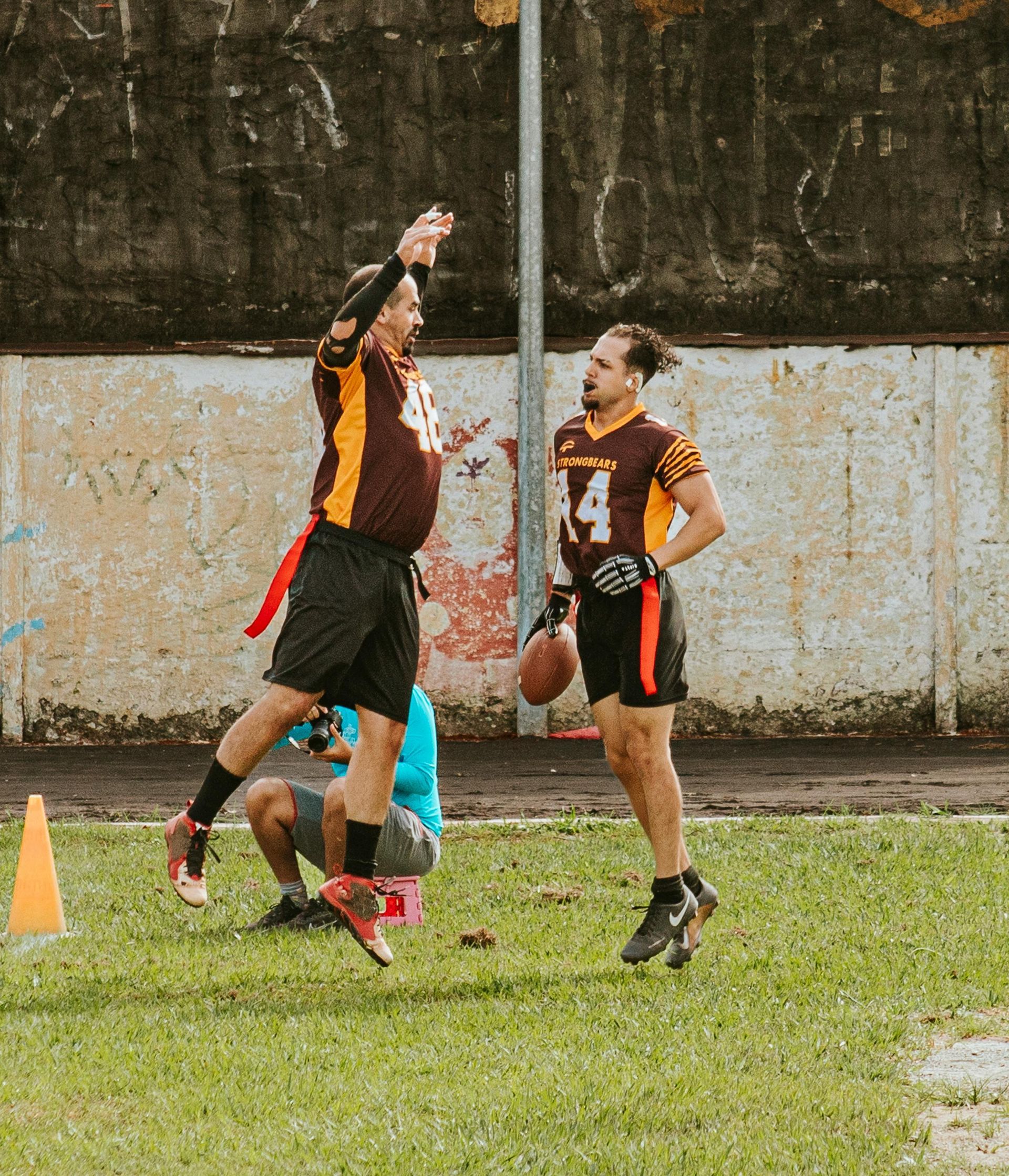 Two athletes in black and orange jerseys jump to catch a football on a wet grassy field.