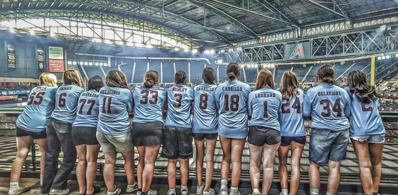 Baseball team in light blue jerseys lined up on the field, facing away under a stadium roof.