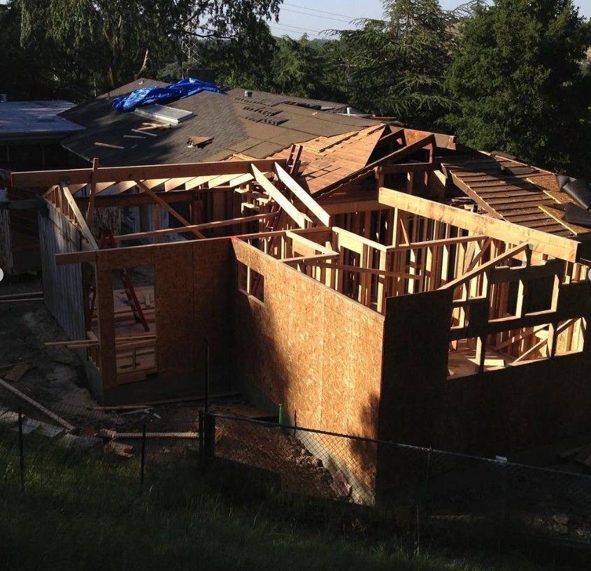 Partially collapsed house with exposed wooden framing and a damaged roof in a wooded yard