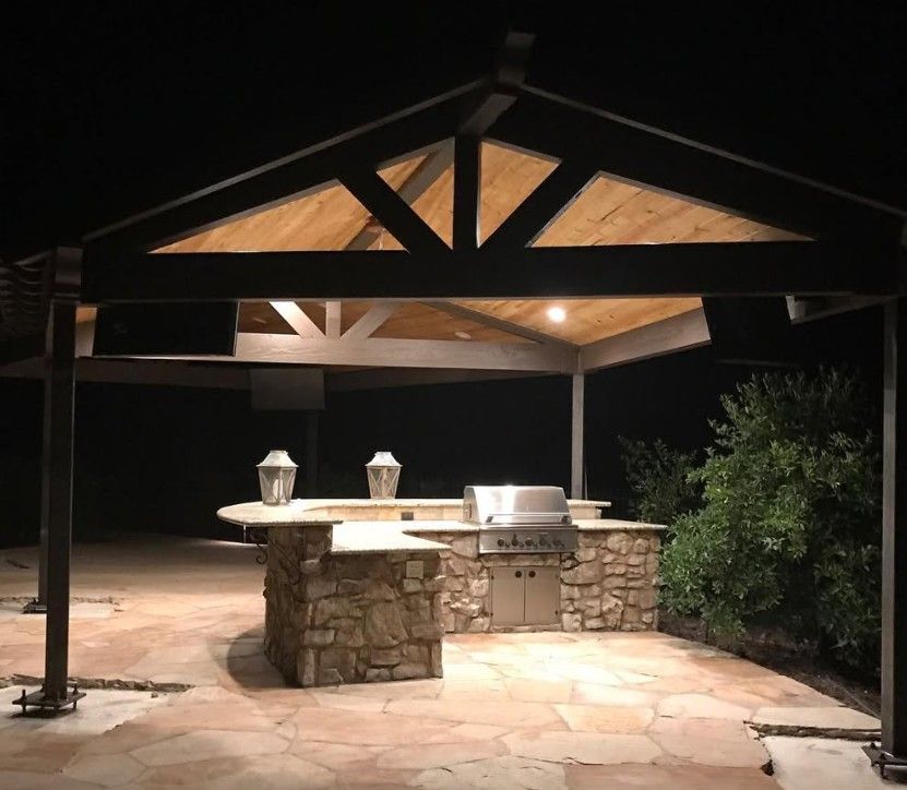 Nighttime outdoor stone grill under a wooden pergola, lit by warm lights on a patio.