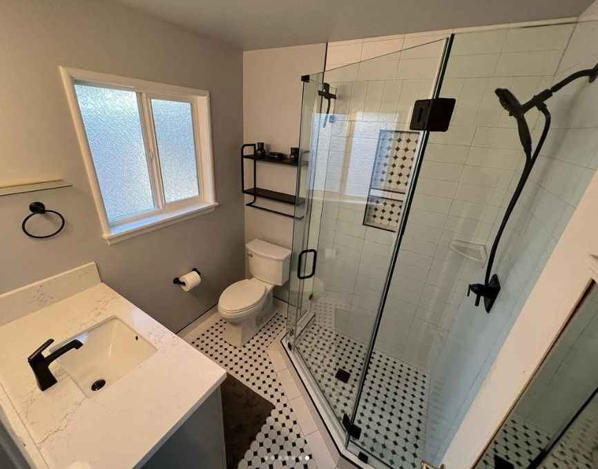 Modern bathroom with walk-in glass shower, toilet, sink, window, and black-and-white tile floor.