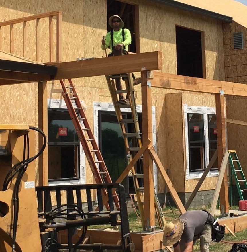Workers building a wooden house frame, with one person on a ladder and another crouching below.