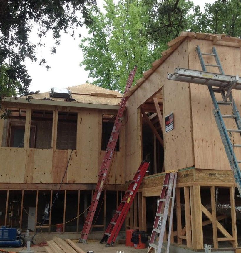 House under construction with ladders and exposed framing, surrounded by trees.