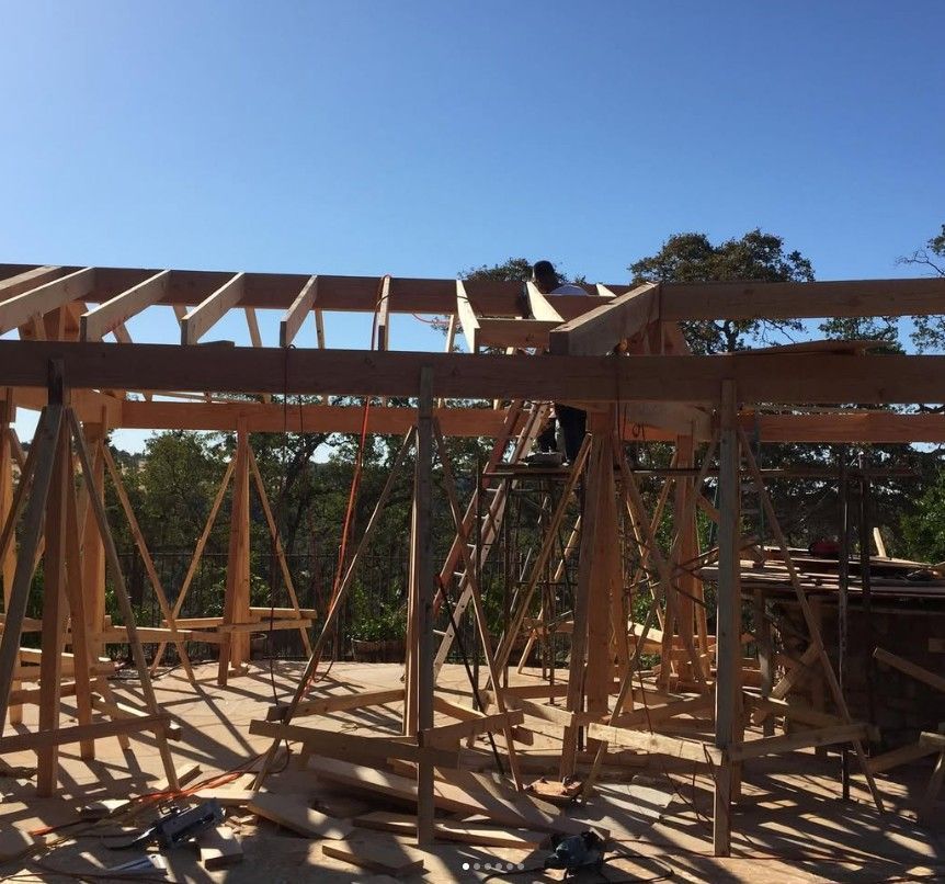 Wooden house frame under construction on a sunny day, with roof trusses and scaffolding.