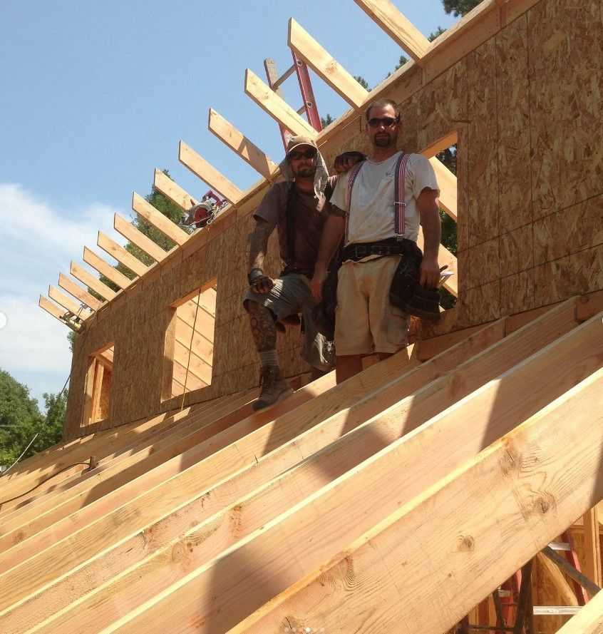 Two workers stand on a wooden roof frame under construction, with plywood walls and exposed rafters.