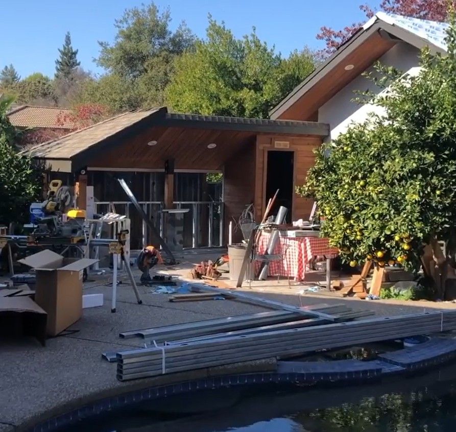 Backyard patio under construction beside a wooden house, with tools, ladders, and stacked lumber.