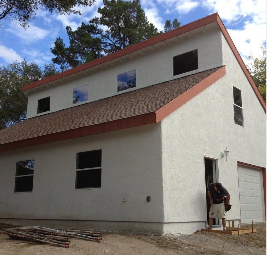 Large white house under construction with red trim, several windows, and a person standing by the garage door.