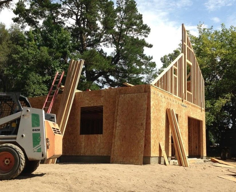 Partially built wooden house framing with ladders and construction equipment outdoors amid trees