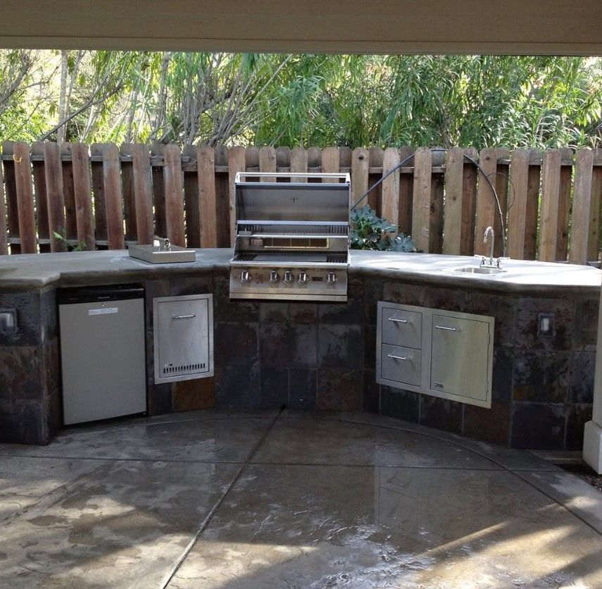 Outdoor built-in stone patio kitchen with grill, sink, cabinets, and small refrigerator under a wooden fence.