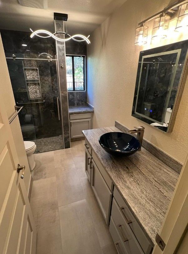 Narrow bathroom with vanity, black bowl sink, large mirror, glass shower, and window at the end.