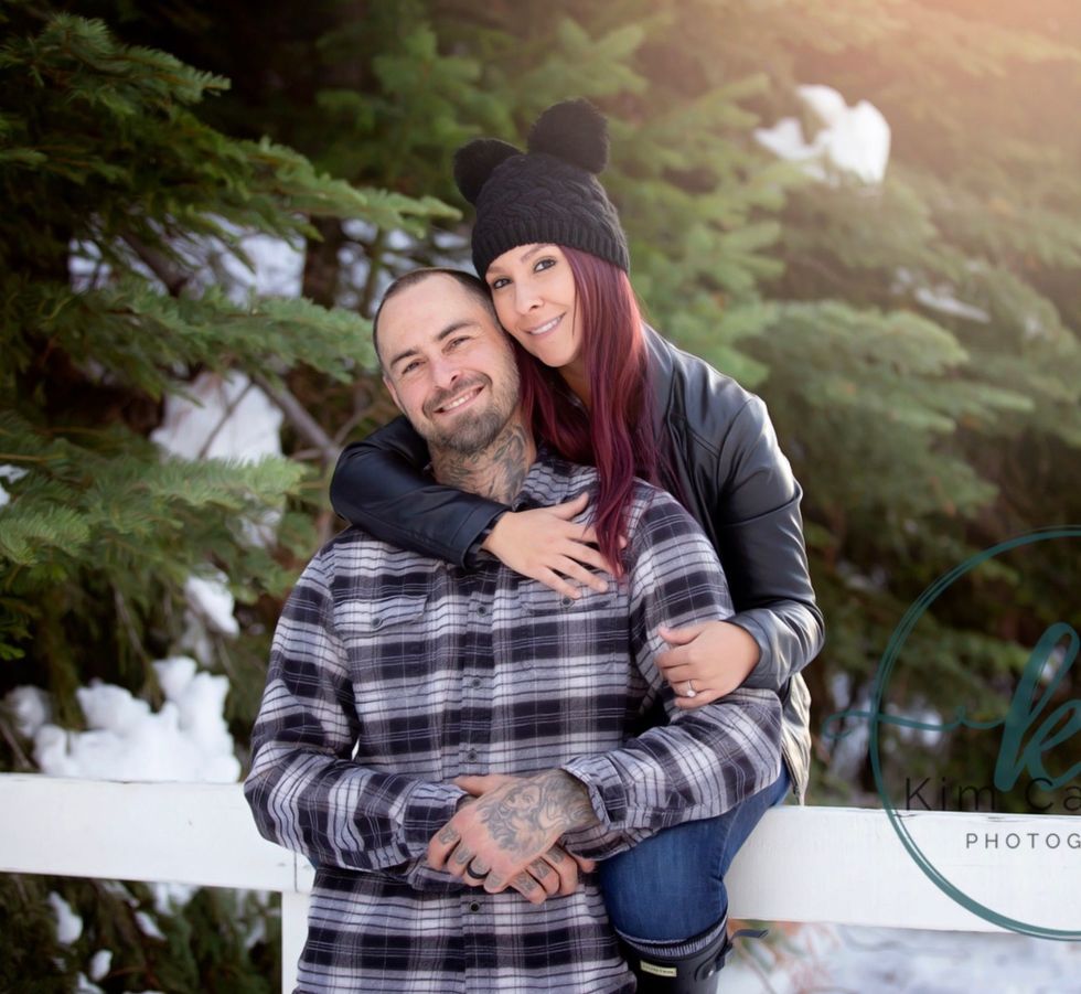Smiling couple in winter clothing embracing outdoors in front of snow and evergreens