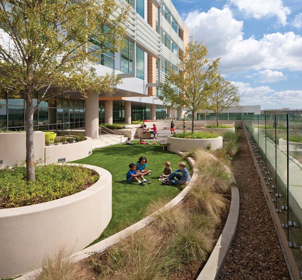 Children sit on grass in a courtyard with trees, shrubs, and a modern building.