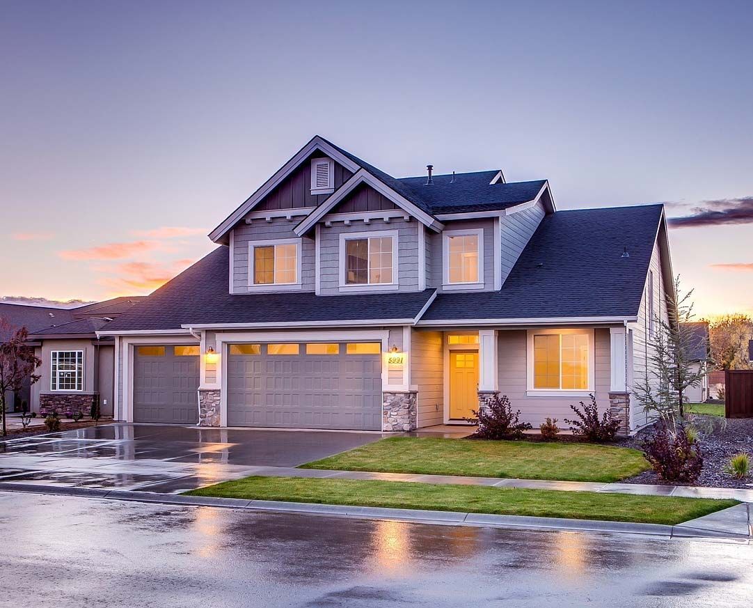 Two-story house with gray siding, blue roof, two-car garage, and wet driveway at dusk.