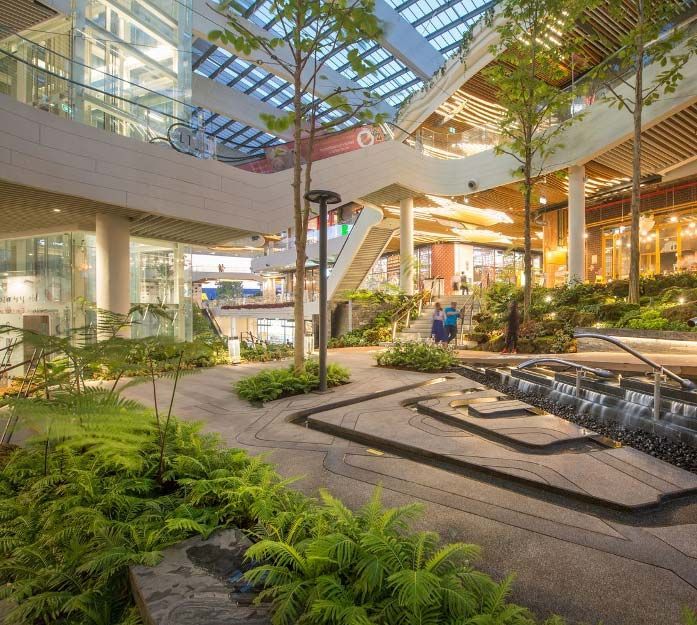 Modern shopping mall interior with greenery, walkways, and cascading water features.