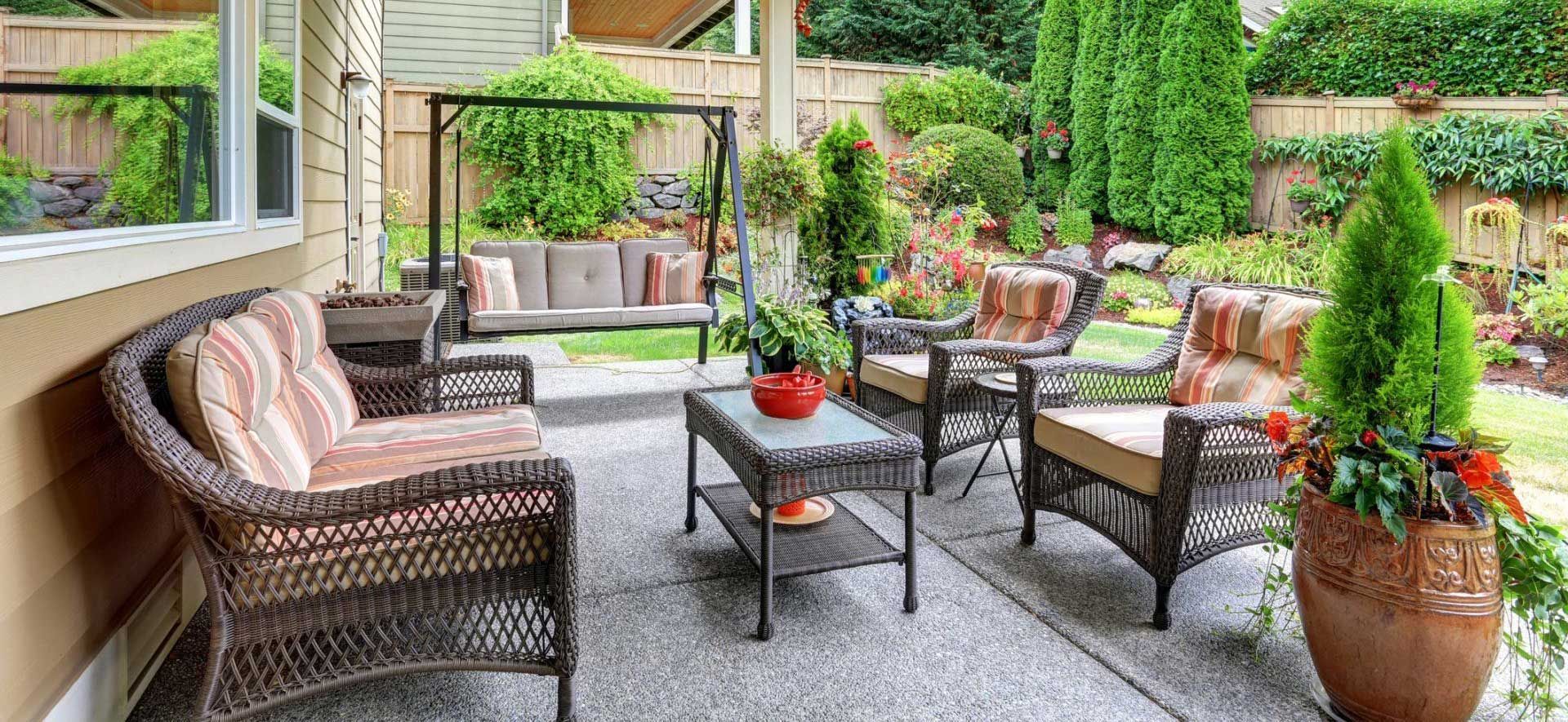 Patio with wicker furniture, swing, table, and potted plants surrounded by lush greenery.