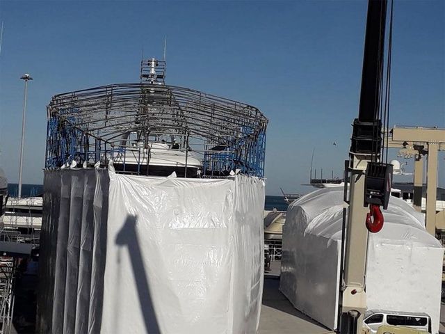 Strutture industriali coperte su un molo, tra cui una struttura metallica blu e una gru sotto un cielo sereno.