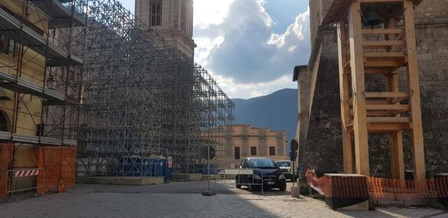 Cortile con un edificio impalcato, un'auto parcheggiata e un cielo nuvoloso tra antiche strutture in pietra.
