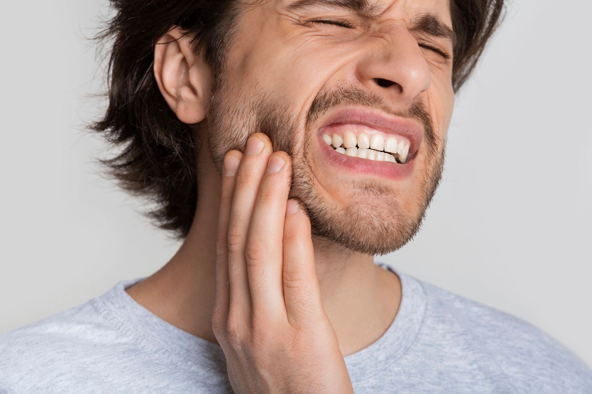 Un uomo con un'espressione sofferente si tiene la mano sulla guancia, a indicare mal di denti o dolore alla mascella, su uno sfondo neutro.