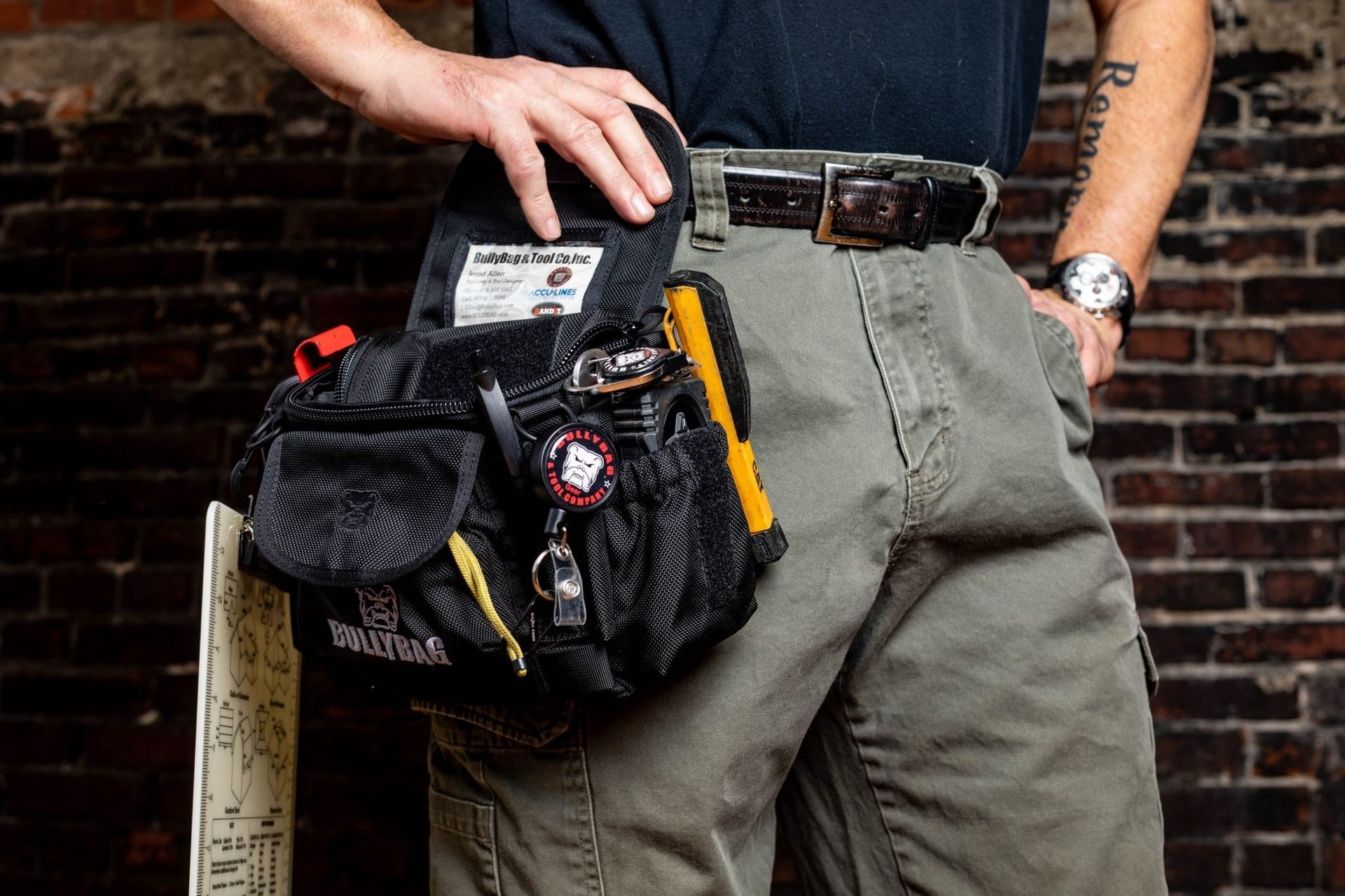 Person wearing tool belt, hand on it, in front of a brick wall.