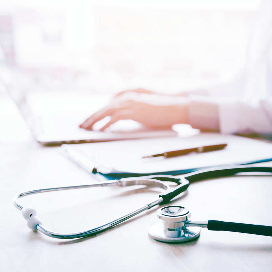Locum doctor's hand on a desk with a laptop and stethoscope.