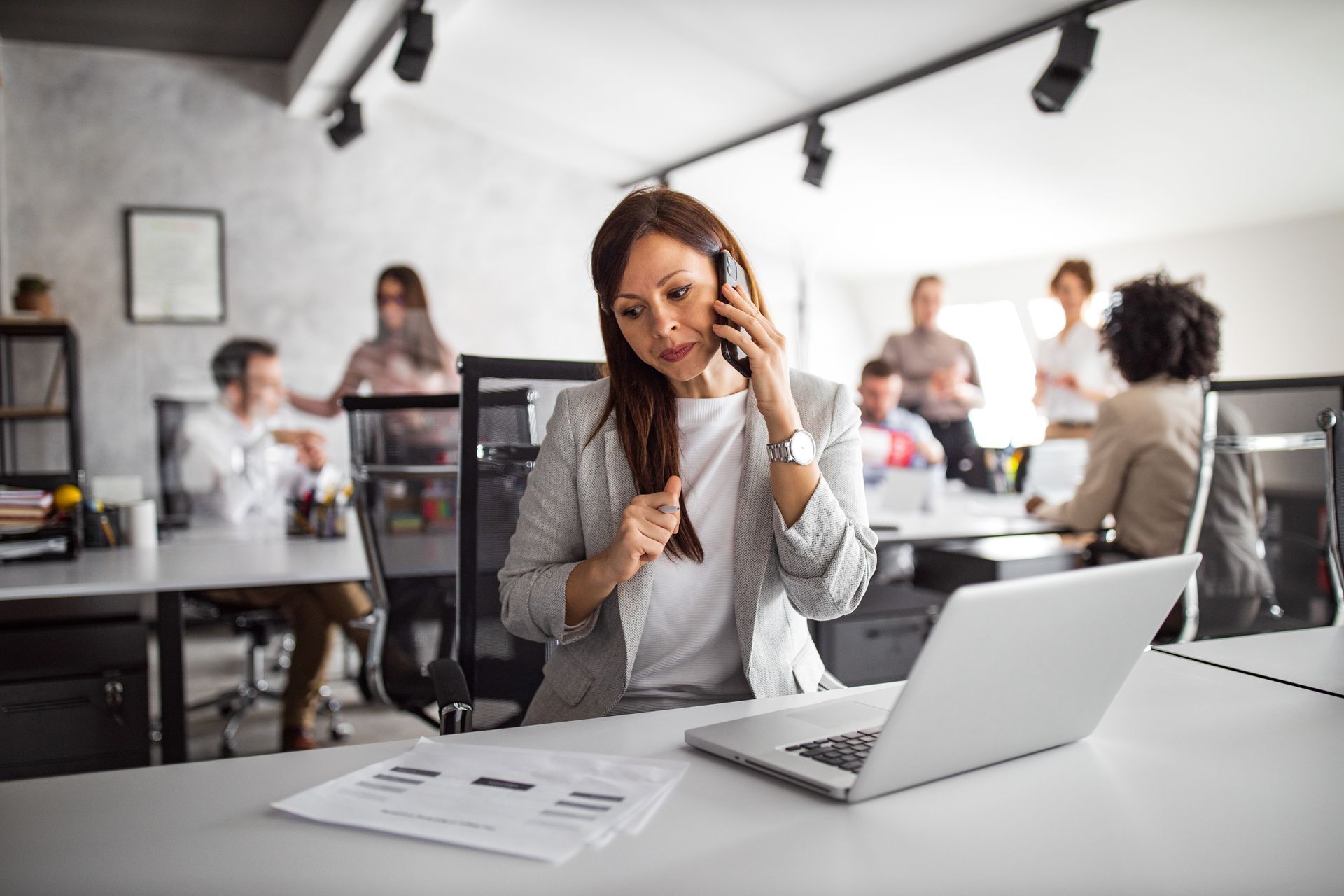 Woman on phone in bright office, working at a desk with a laptop and papers while colleagues meet in background