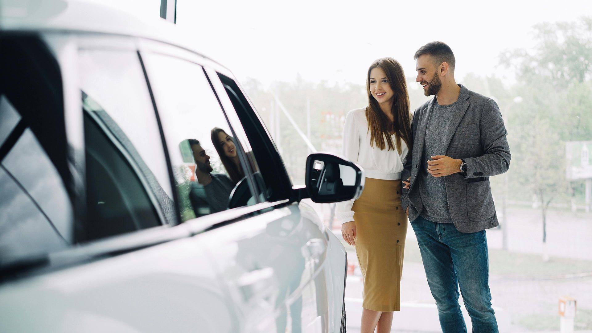 Two people standing beside a white car, looking at the side mirror outdoors in a bright setting