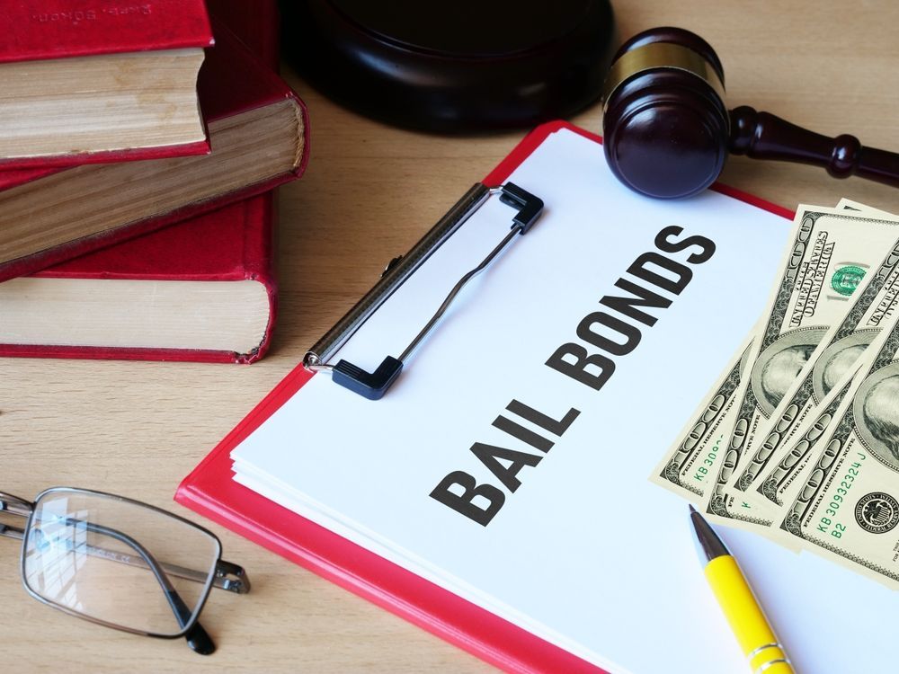 Clipboard labeled “BAIL BONDS” with cash, gavel, glasses, and books on a desk