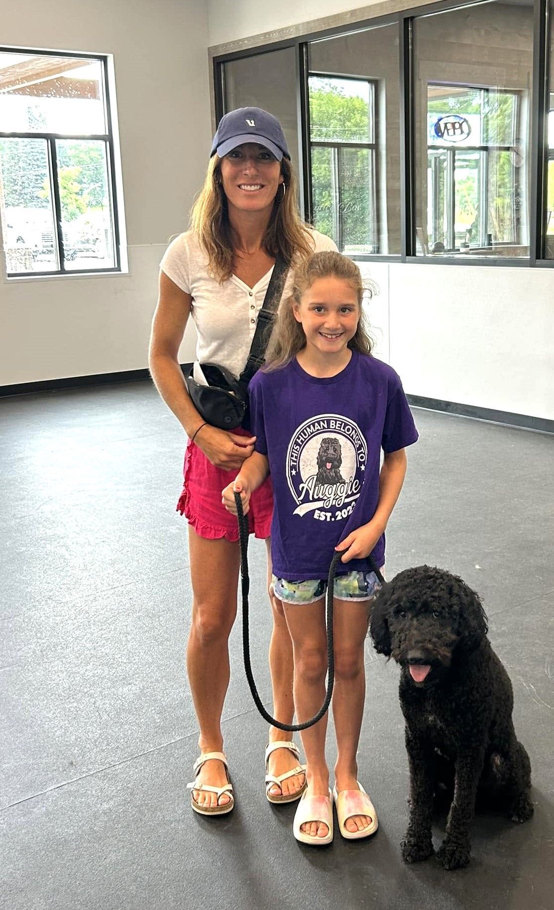 Woman and child standing indoors with a black dog on a leash, smiling in a bright room.
