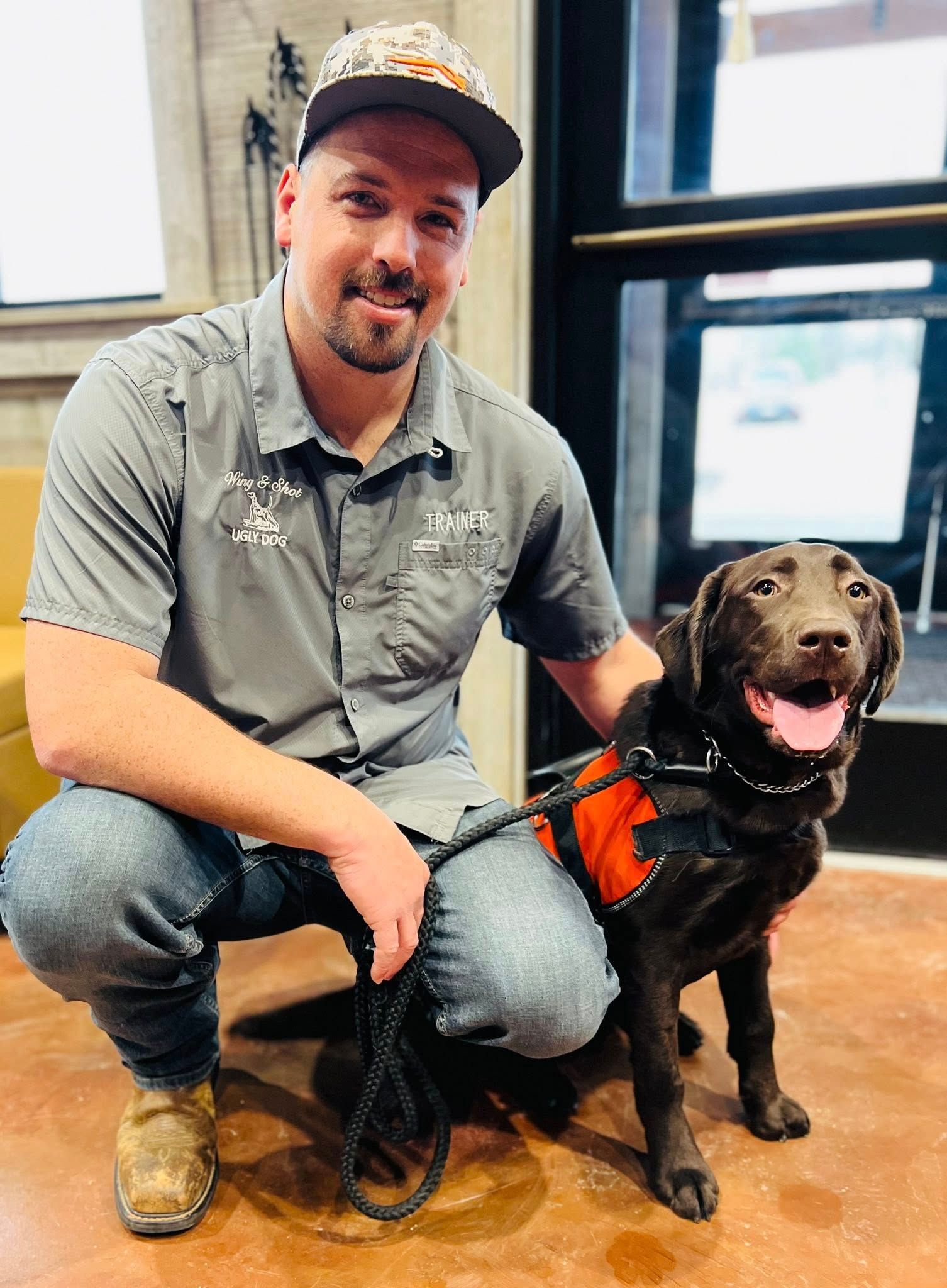 Man kneeling beside a black dog in an orange harness indoors, both looking at the camera.