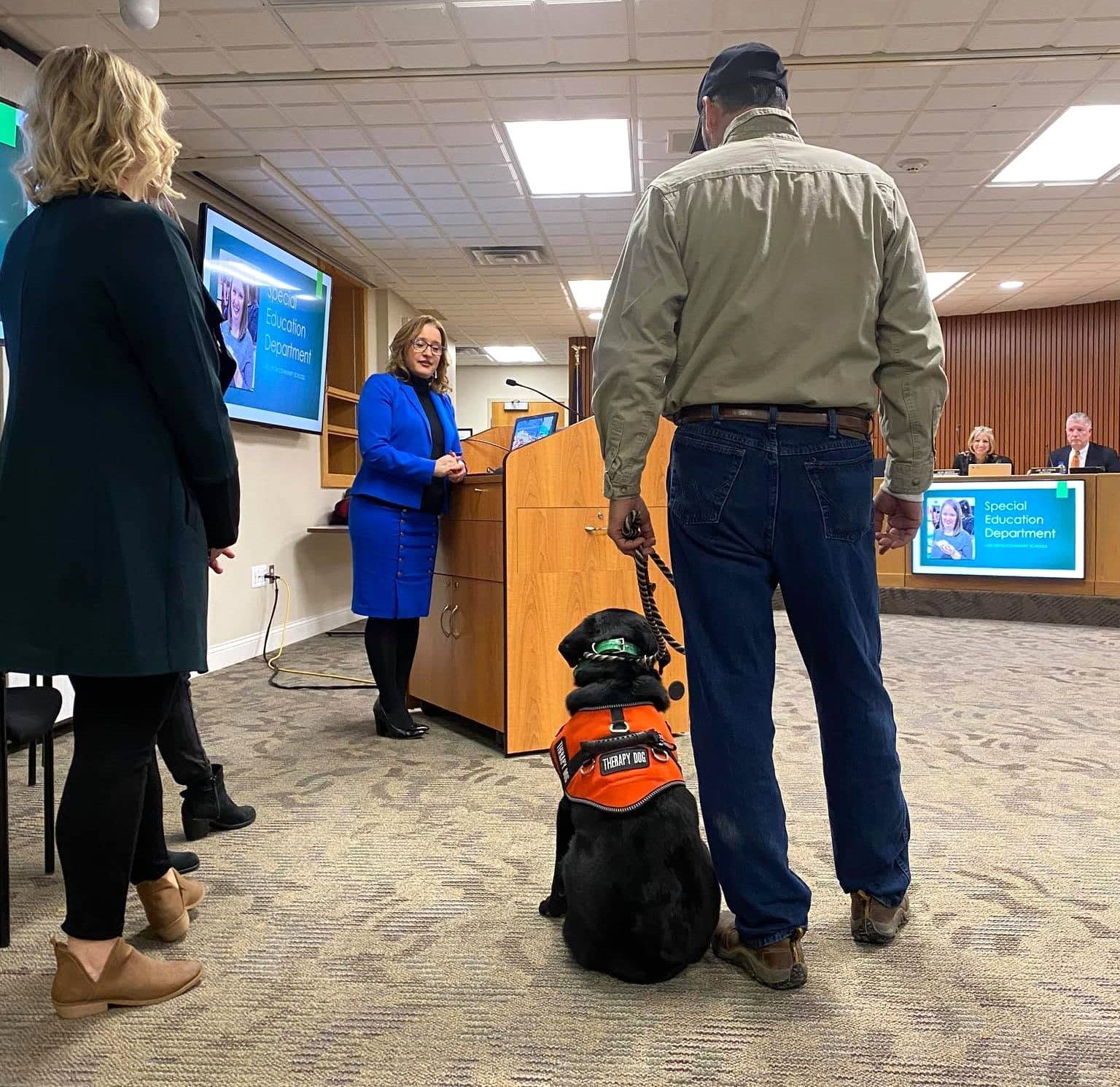 Dog in orange service vest sits beside handler at a podium in a conference room.