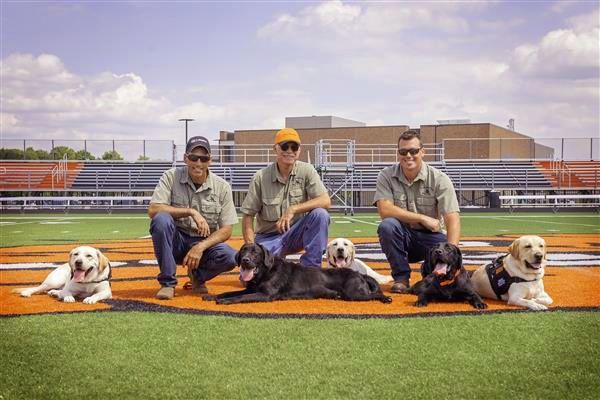 Three people and four dogs on a baseball field, posing on the pitcher's mound under a cloudy sky.