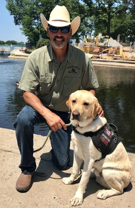 Man in a cowboy hat kneels beside a yellow Labrador by the water with boats and trees in the background