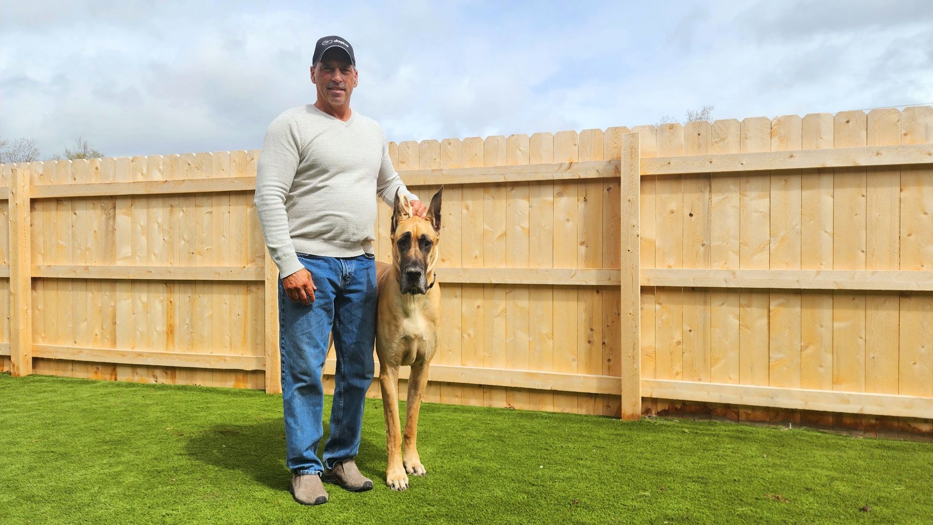 Man standing on grass beside a tan dog in front of a wooden fence.