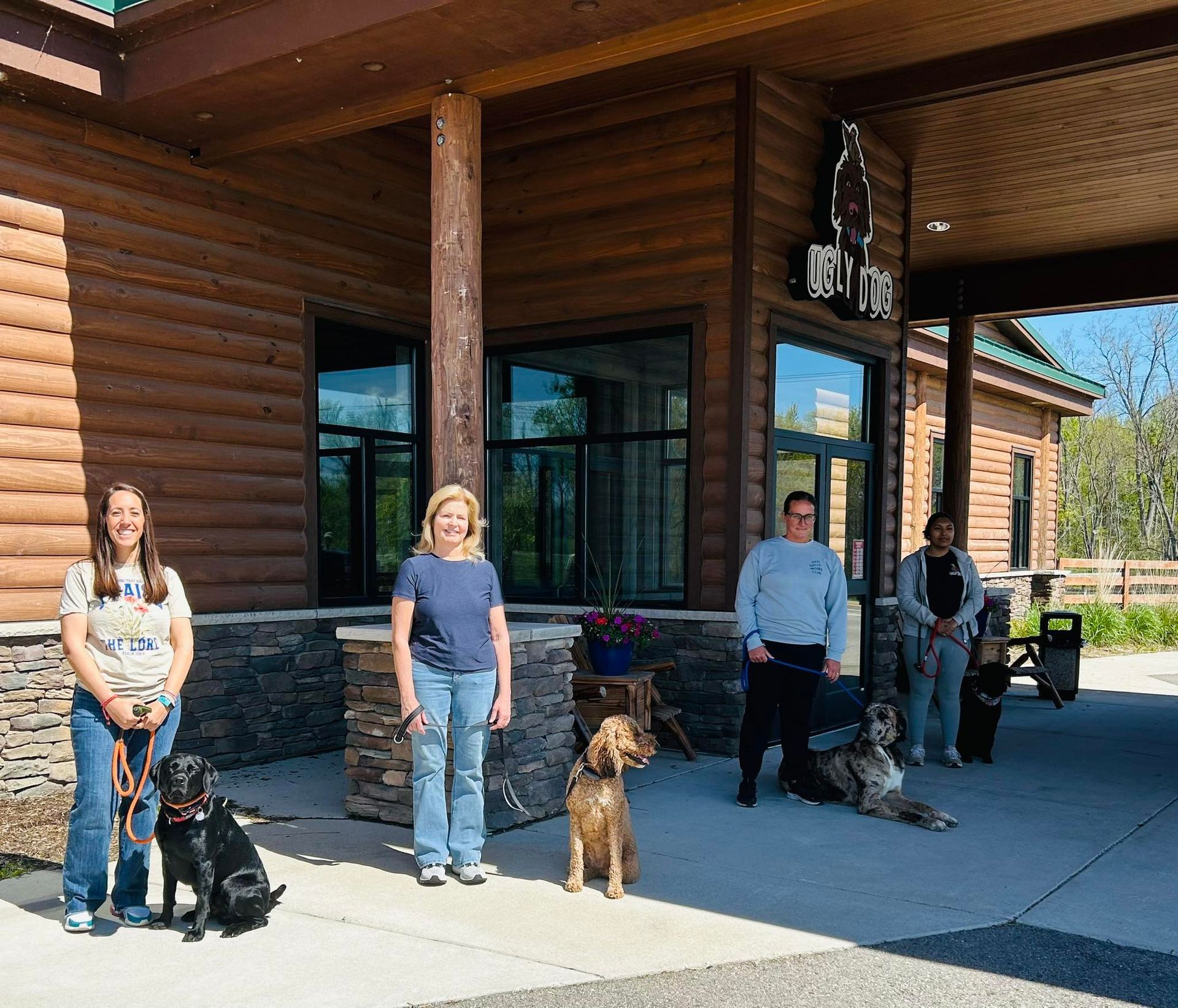 Four people with dogs stand outside a wooden building in bright sunlight.