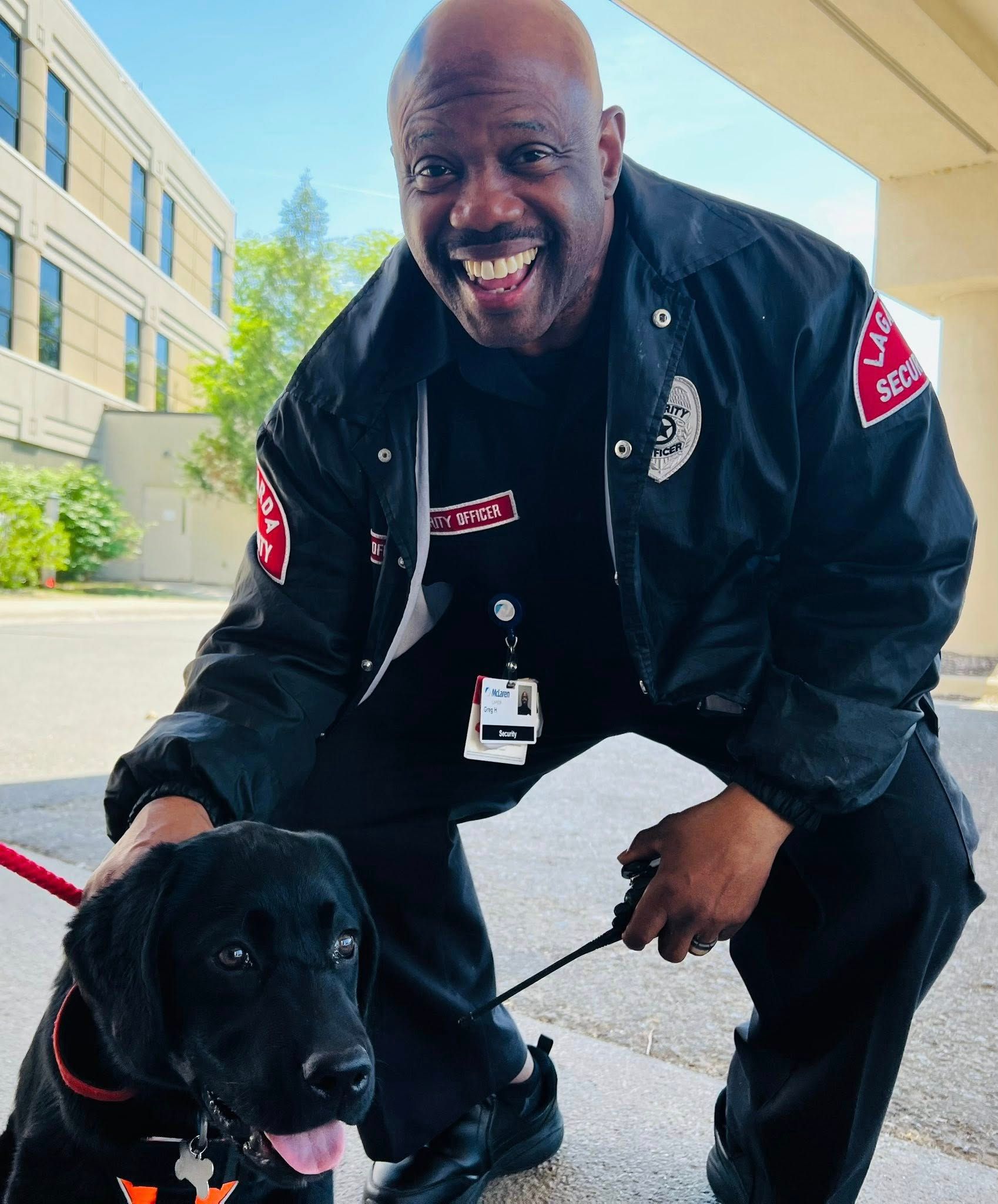 Black dog with a handler in a black uniform crouching and smiling outdoors by a building.