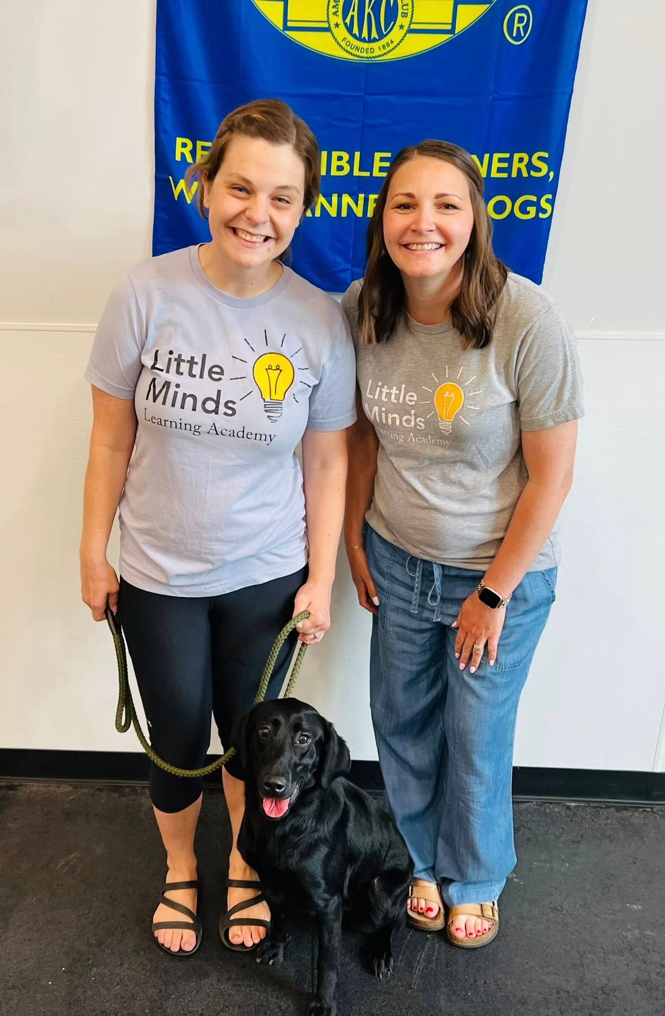 Two women smiling beside a black dog in front of a blue banner indoors.