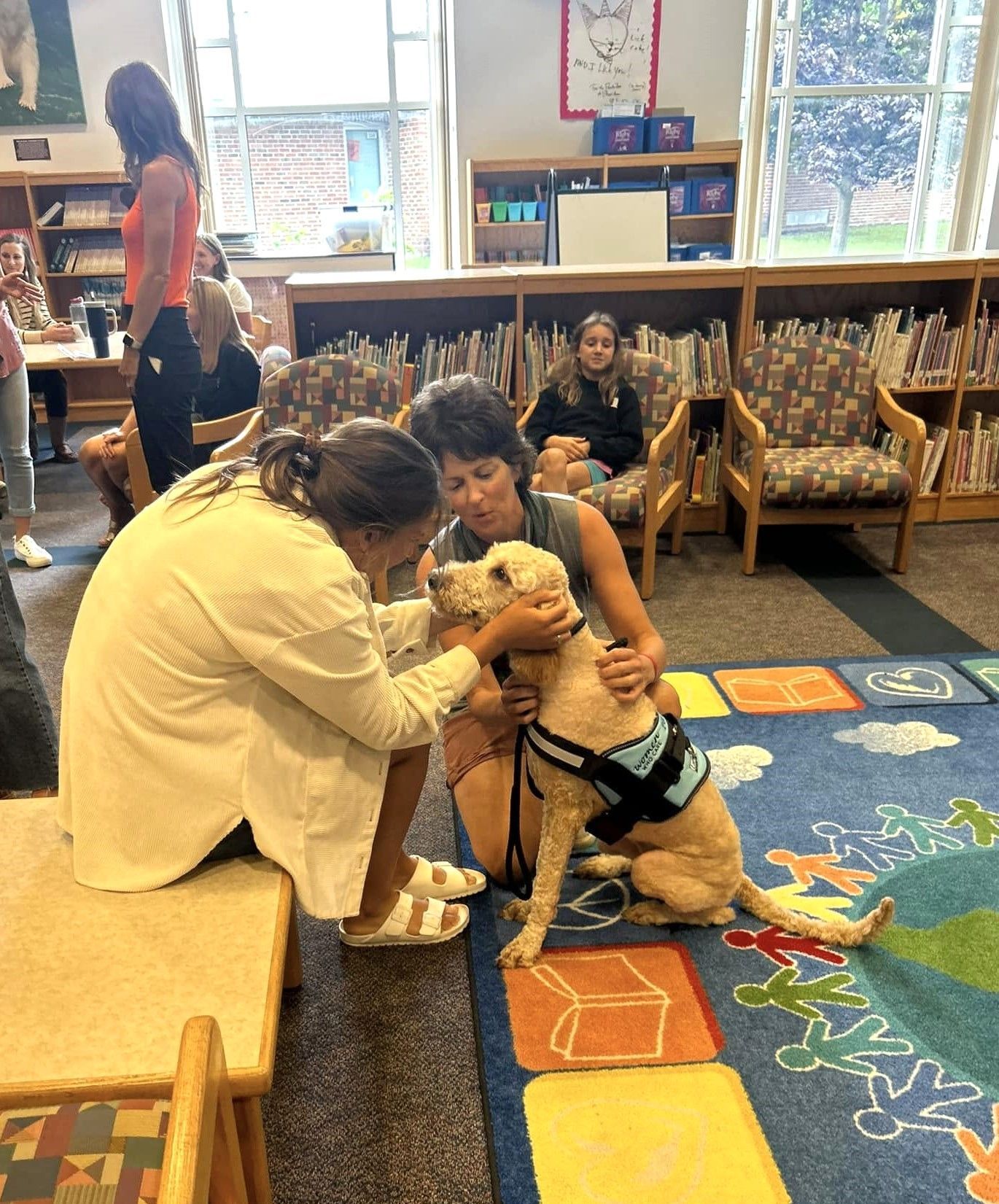 Adult comforting a dog in a classroom with colorful carpet and chairs while children watch nearby