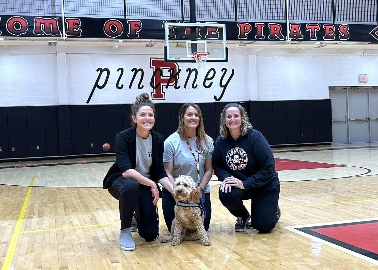 Three people kneel with a dog on a gym floor in front of a basketball hoop and school banner.