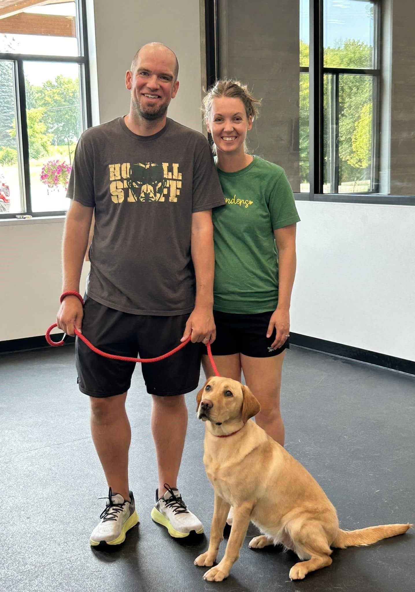 Two people standing with a tan dog inside a gym, one holding a red leash