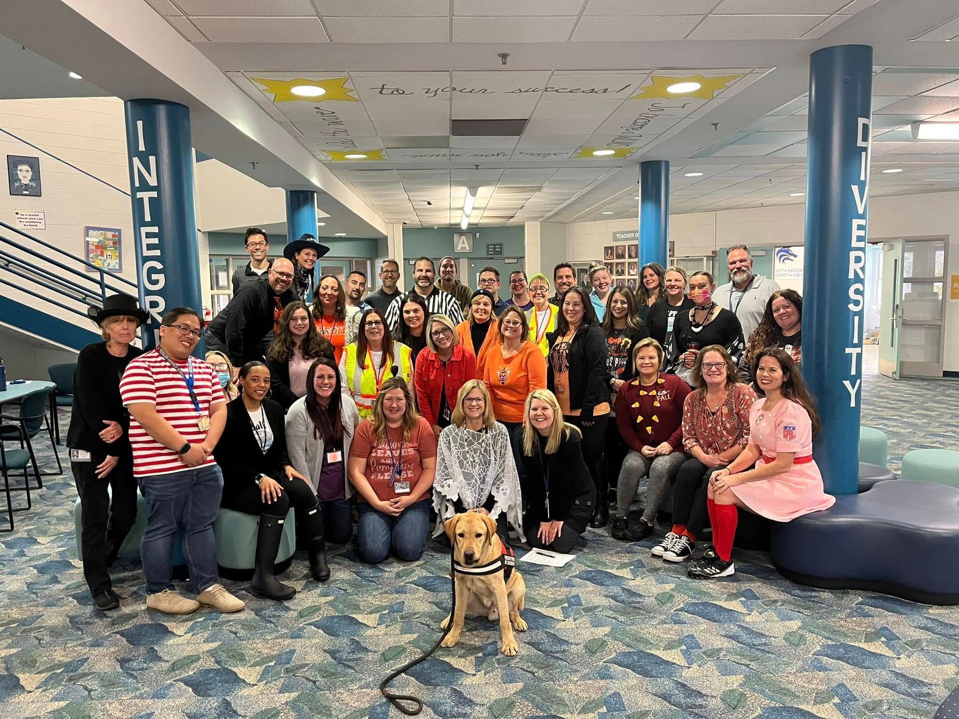 Large group posing in a university lobby, with a service dog in front and blue columns labeled “INTEGRITY” and “VERSITY”