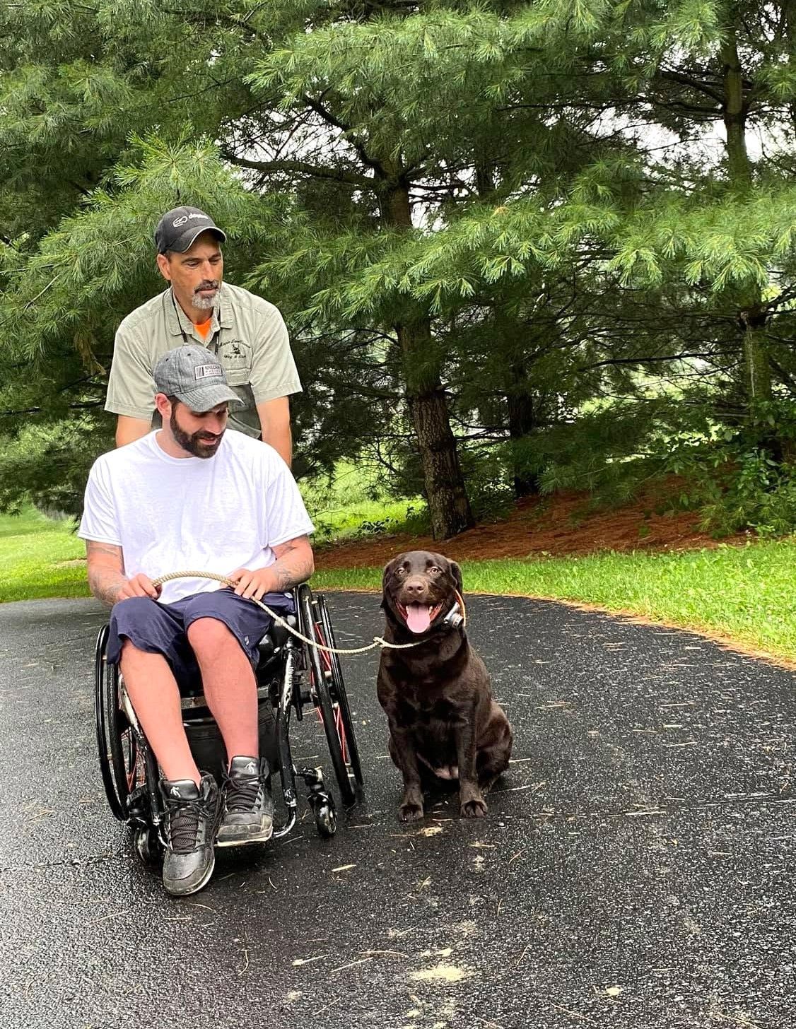 Man in a wheelchair on a gravel path with a dog, another man standing behind, trees in the background