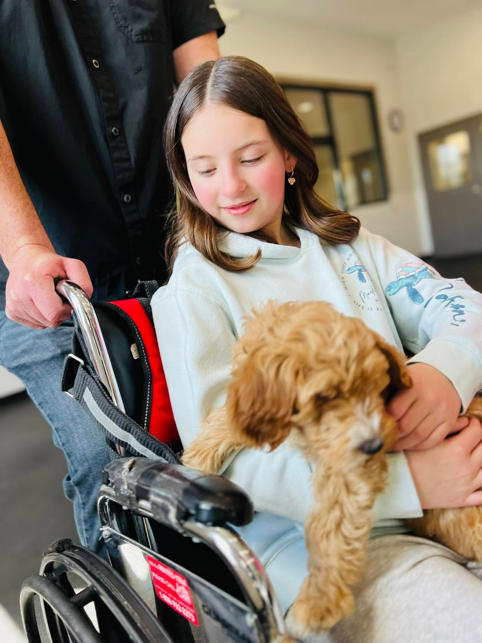 Girl in a wheelchair holding a curly dog, with an adult standing beside her indoors.