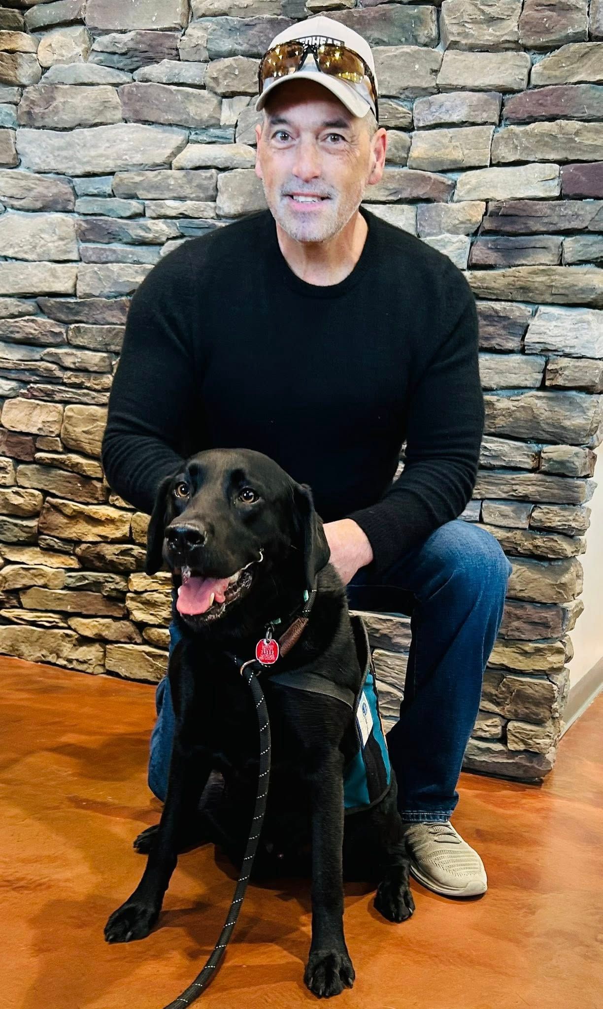 Man kneels behind black dog in a cap, posing indoors in front of a stone wall.