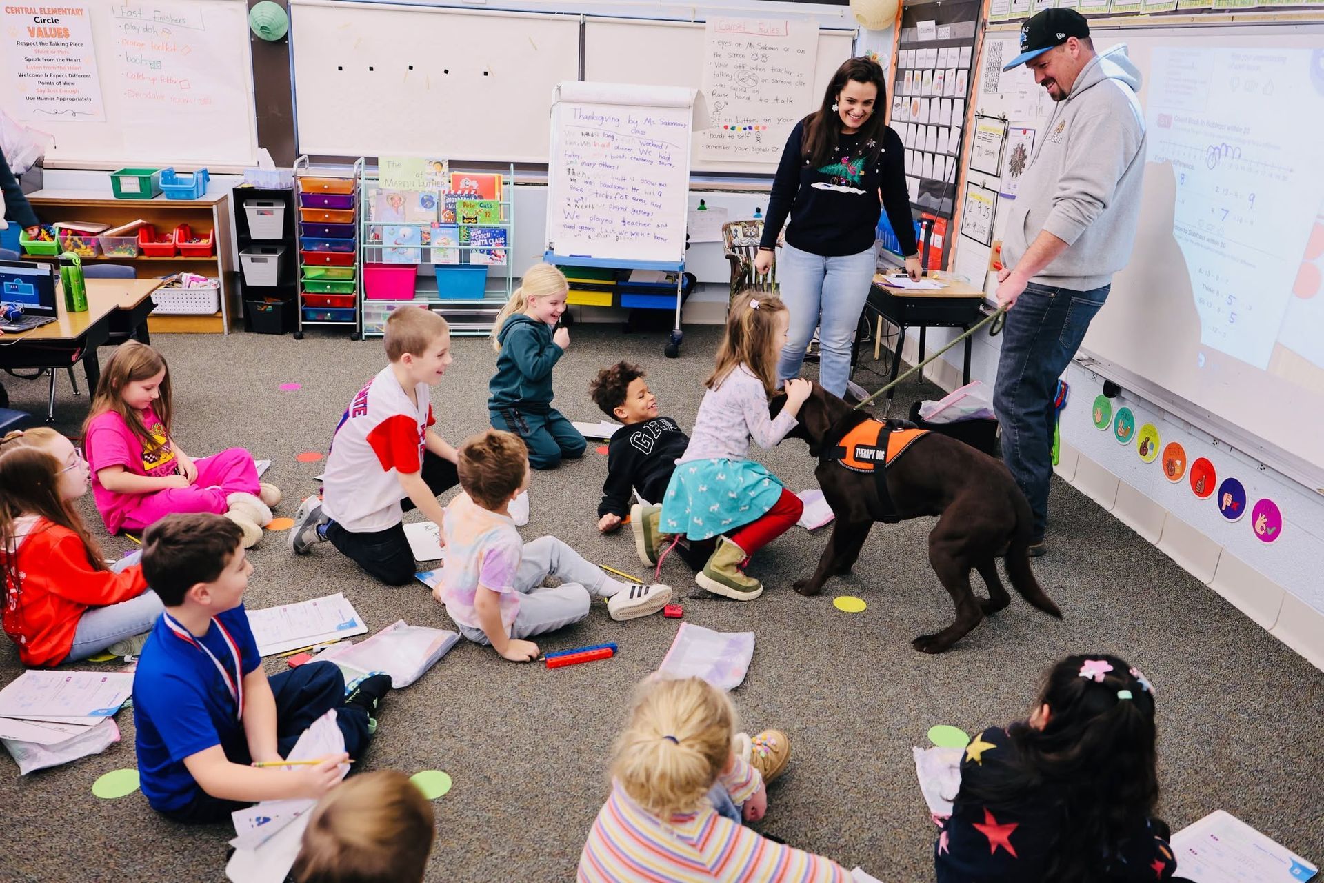 Teacher and students sit on the floor with a black dog in a colorful classroom.